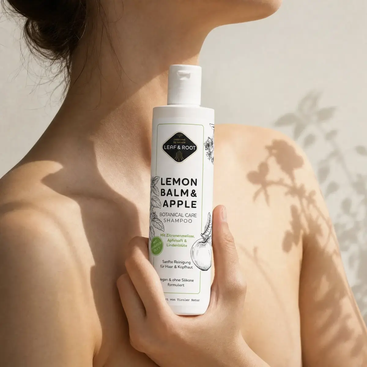 Frau hält die Shampoo-Flasche vor Schulter und Schlüsselbein, Schatten eines Melissenzweigs auf der Haut