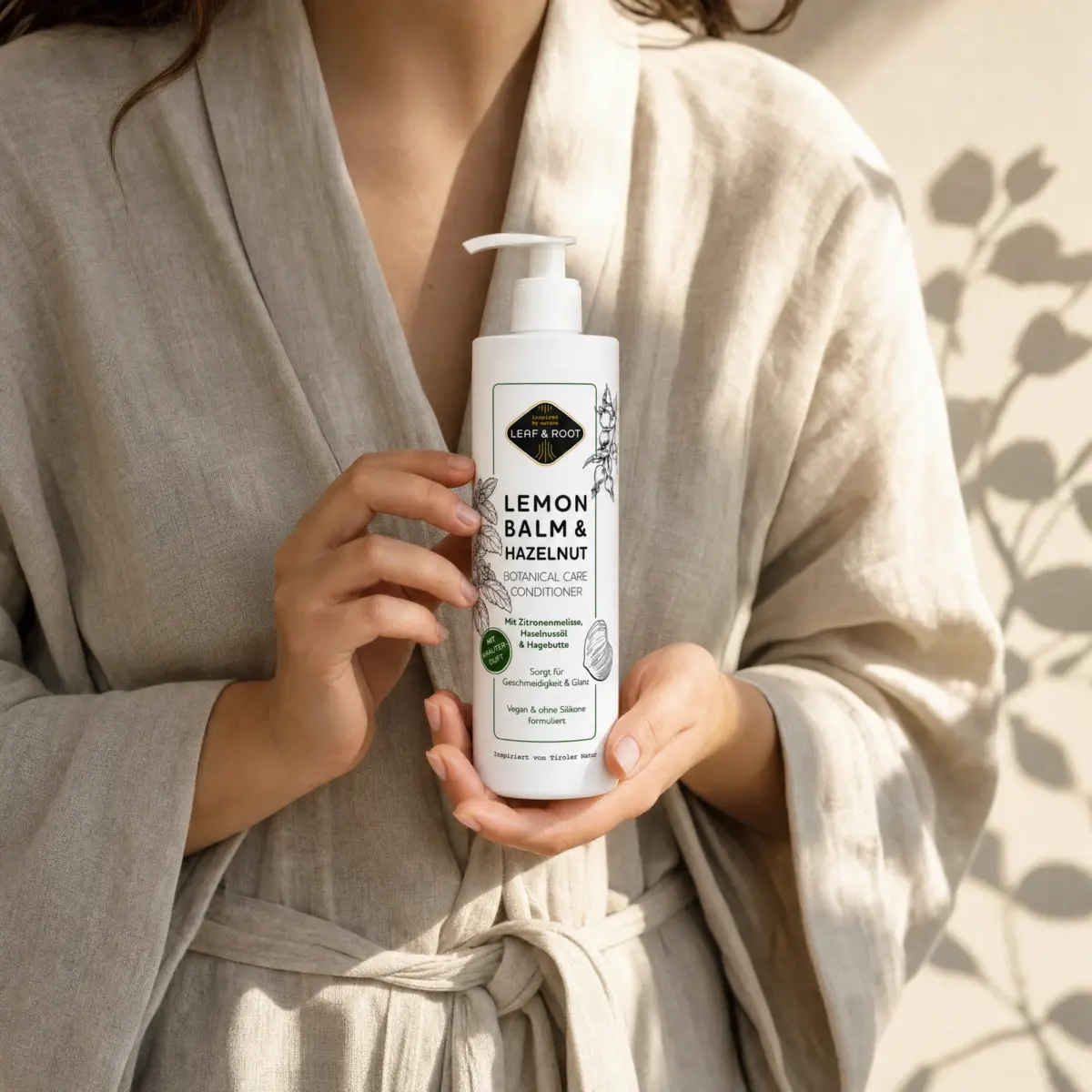 Frau im Leinenmantel hält die Conditioner-Flasche in beiden Händen, Schatten von Blattwerk auf dem Stoff